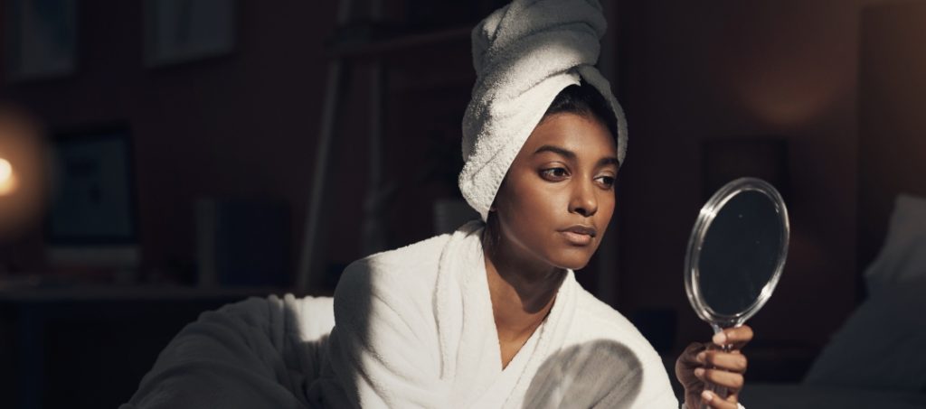 an woman medium-dark brown skin examines her face in a hand mirror